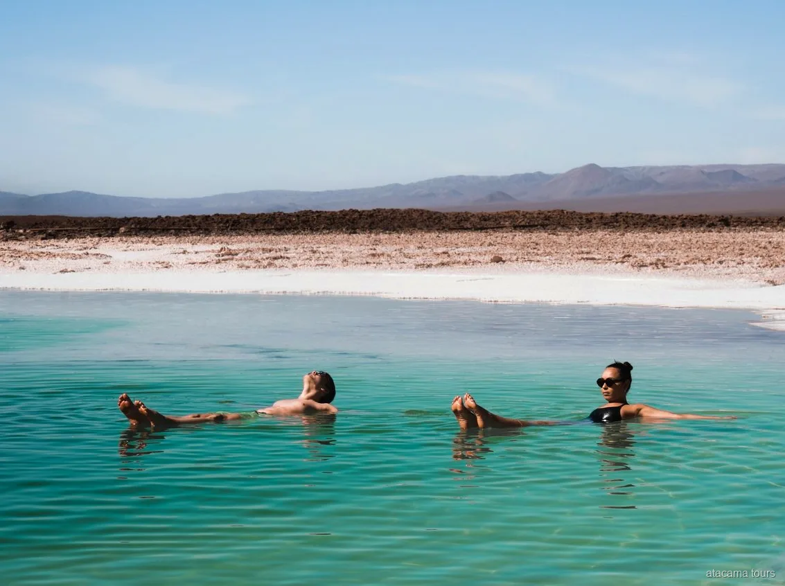 Atacama tours combo, two visitors floating in a turquoise salt lagoon with desert mountains behind