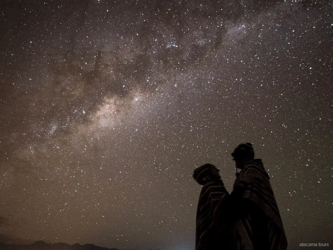 Atacama stargazing tours, two silhouettes wrapped in ponchos beneath a dense band of the Milky Way