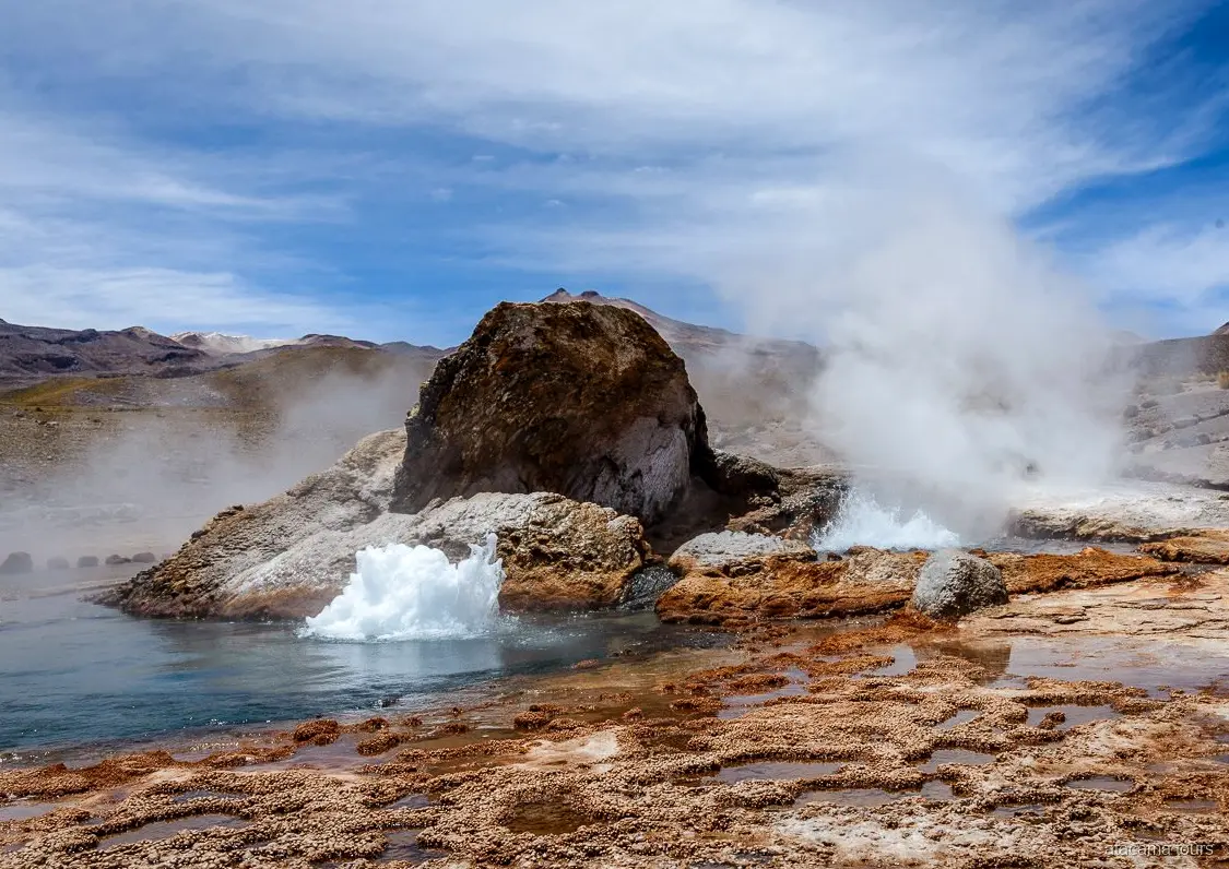 Atacama tours naar El Tatio, borrelende geiser naast een groot rotsblok met stoom en mineraalterrassen