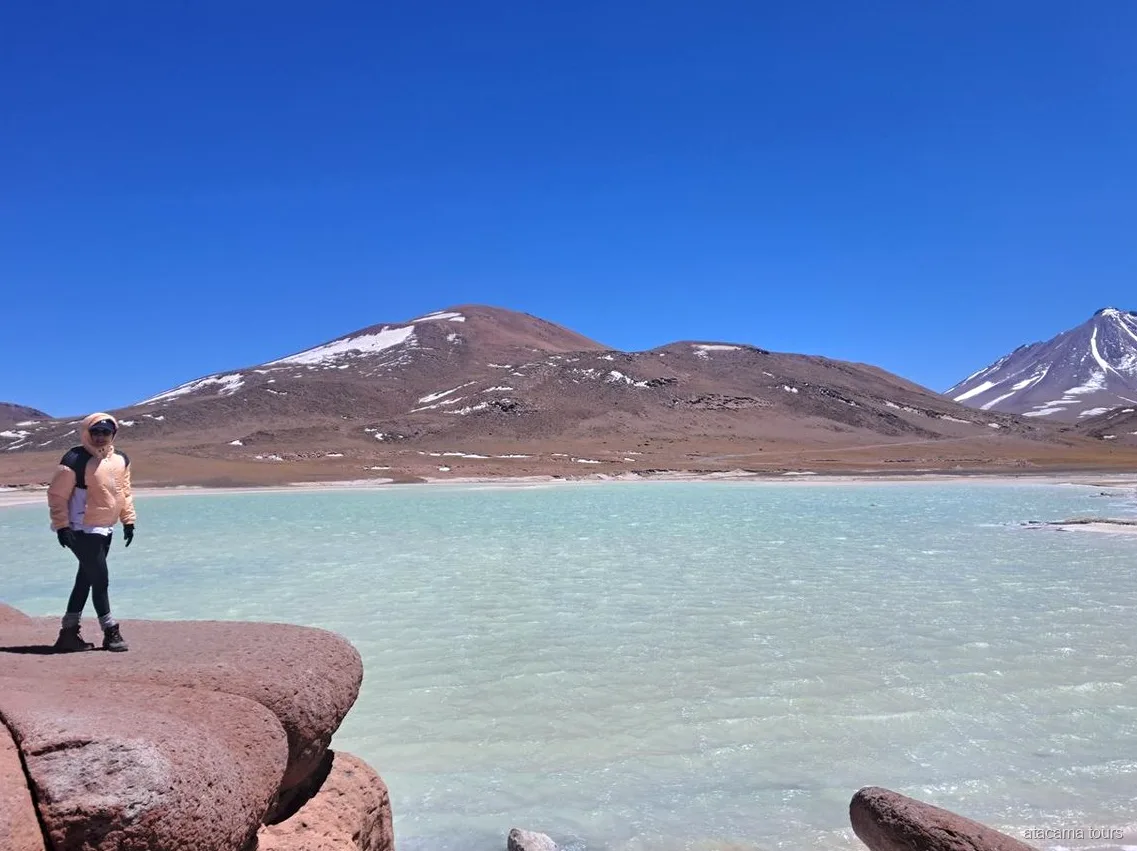 Passeios no Atacama a Piedras Rojas, visitante sobre rochas vermelhas ao lado de uma laguna turquesa