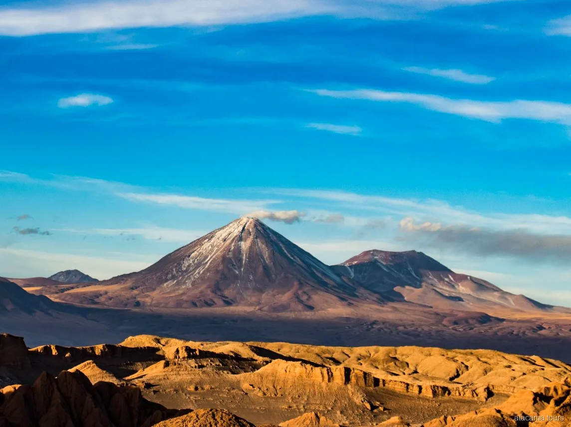 Paesaggio dei tour nel deserto di Atacama, cono vulcanico innevato sopra creste desertiche dorate ed erose