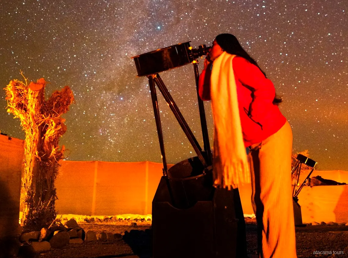 Tours en Atacama, viajera mirando por un gran telescopio bajo el cielo estrellado del desierto de noche