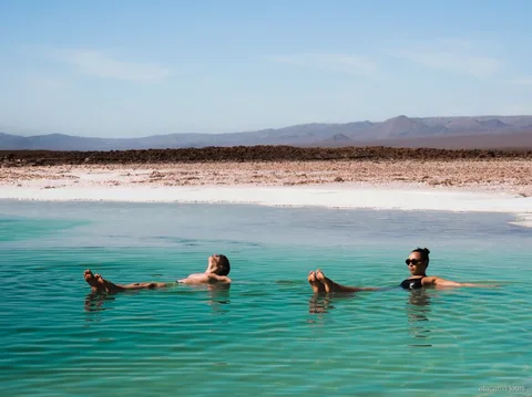Tours en Atacama, dos visitantes flotando en una laguna salada turquesa con montañas del desierto al fondo