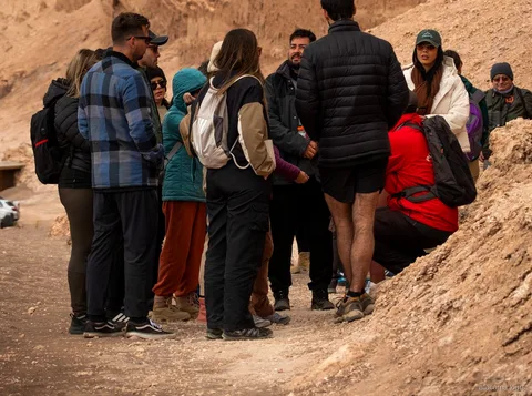 アタカマツアーで歩く月の谷、岩肌に囲まれた砂地のトレイルでガイドの説明に耳を傾ける、ダウンジャケット姿の大勢の参加者グループ